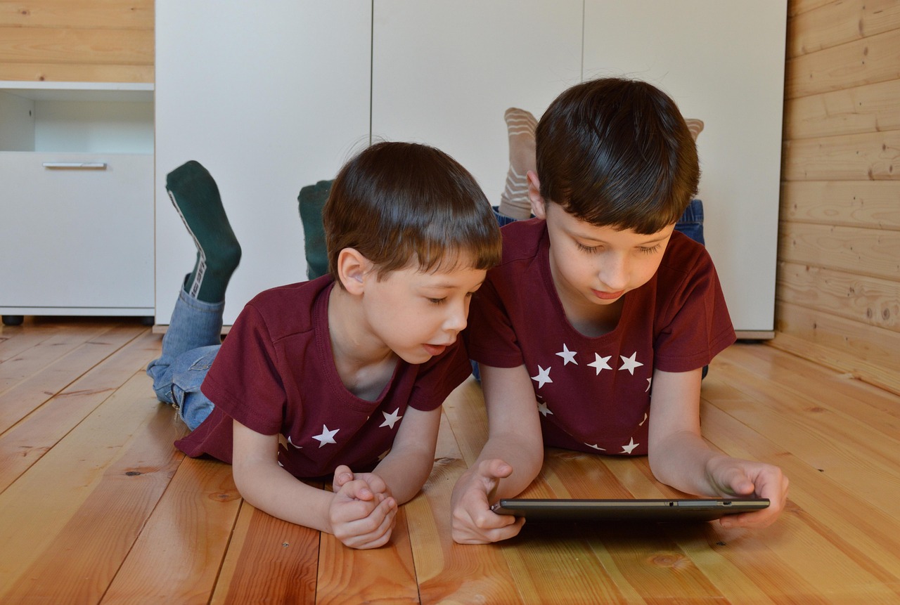 Dos niños que serán hermanos tumbados en el suelo de madera viendo una tablet