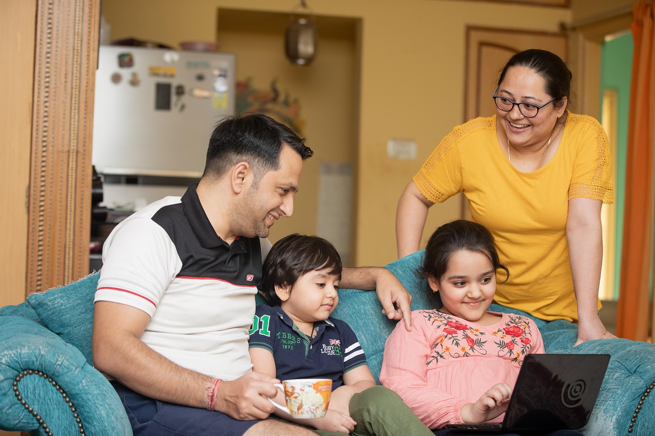 Familia de cuatro integrantes en el salón de su casa donde están sentados en el sillón el padre y los dos hijos. La madre está de pie, todos sonríen