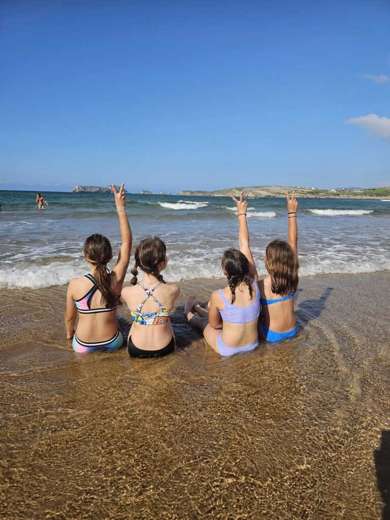 Cuatro niñas de espaldas sentadas en la orilla de la playa con una brazo levantado con el simbolo de victoria