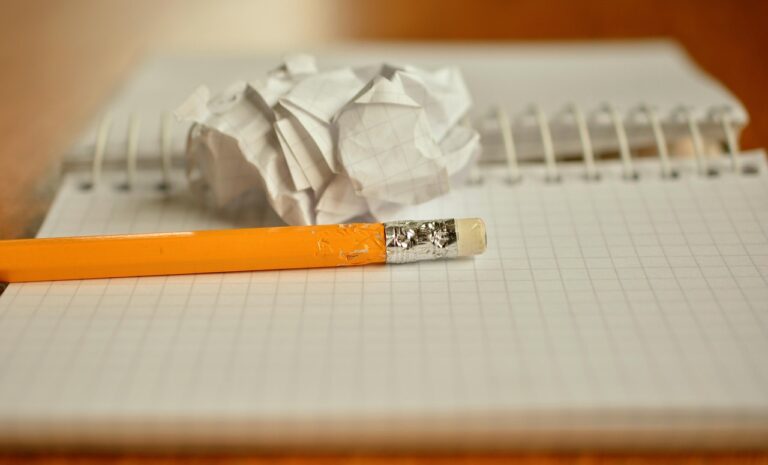 En primer plano se ve un cuaderno abierto, de cuadros con un lápiz amarillo mordido y una bola de papel