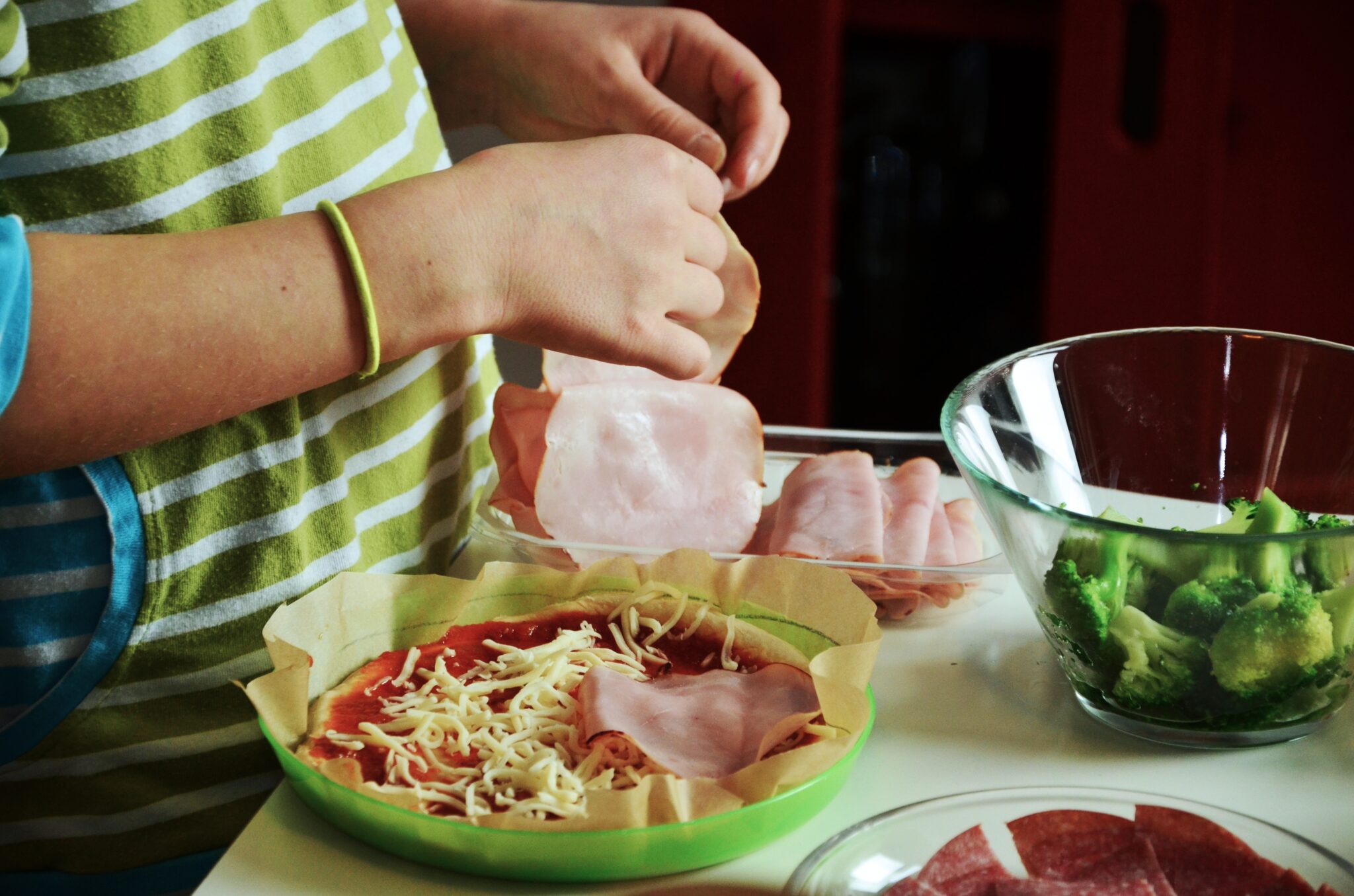 Primer plano de una manos de niño preparando una pizza con queso, jamon y brocoli