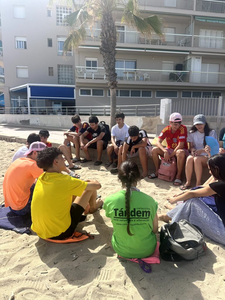grupo de adolescentes sentados en circulo en la playa con su monitora