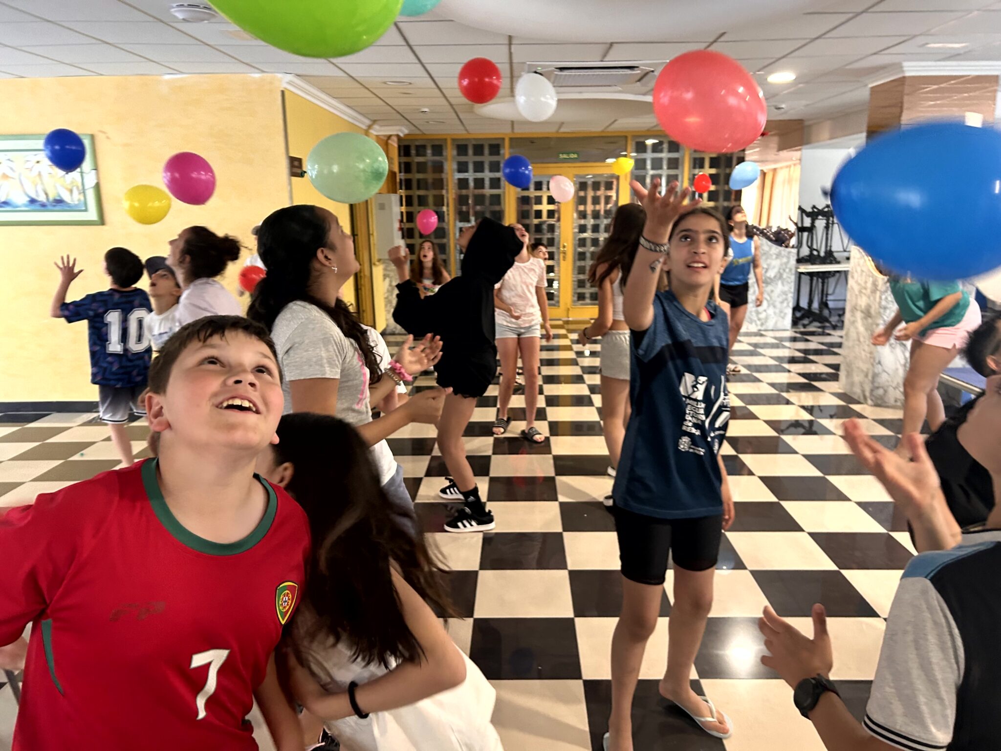 grupo de chavales haciendo una dinámica de globos en una sala con baldosas blancas y negras