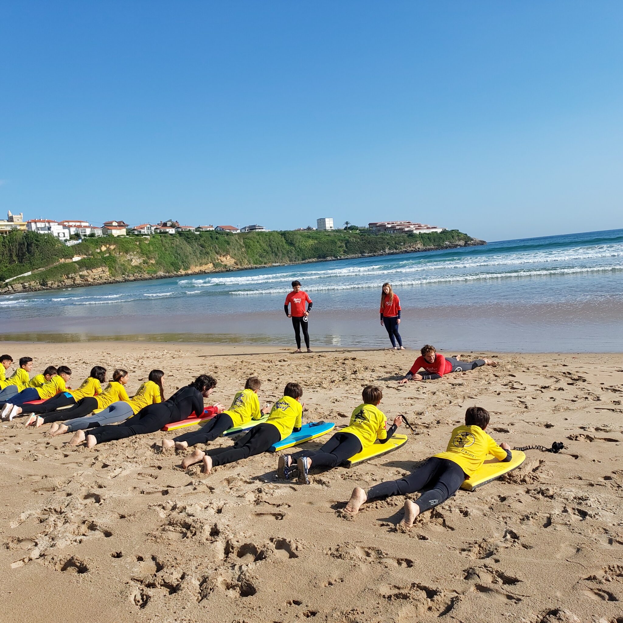 grupo de primaria con neopreno y camiseta amarilla tumbados en la playa sobre tablas de bodysurf escuchando las explicaciones