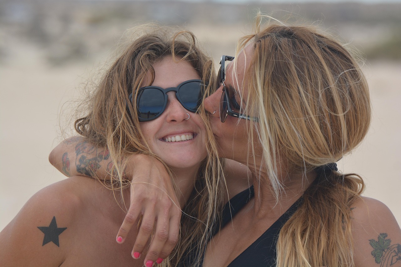 Dos chicas jóvenes en la playa con gafas de sol, una besa a la otra en la mejilla