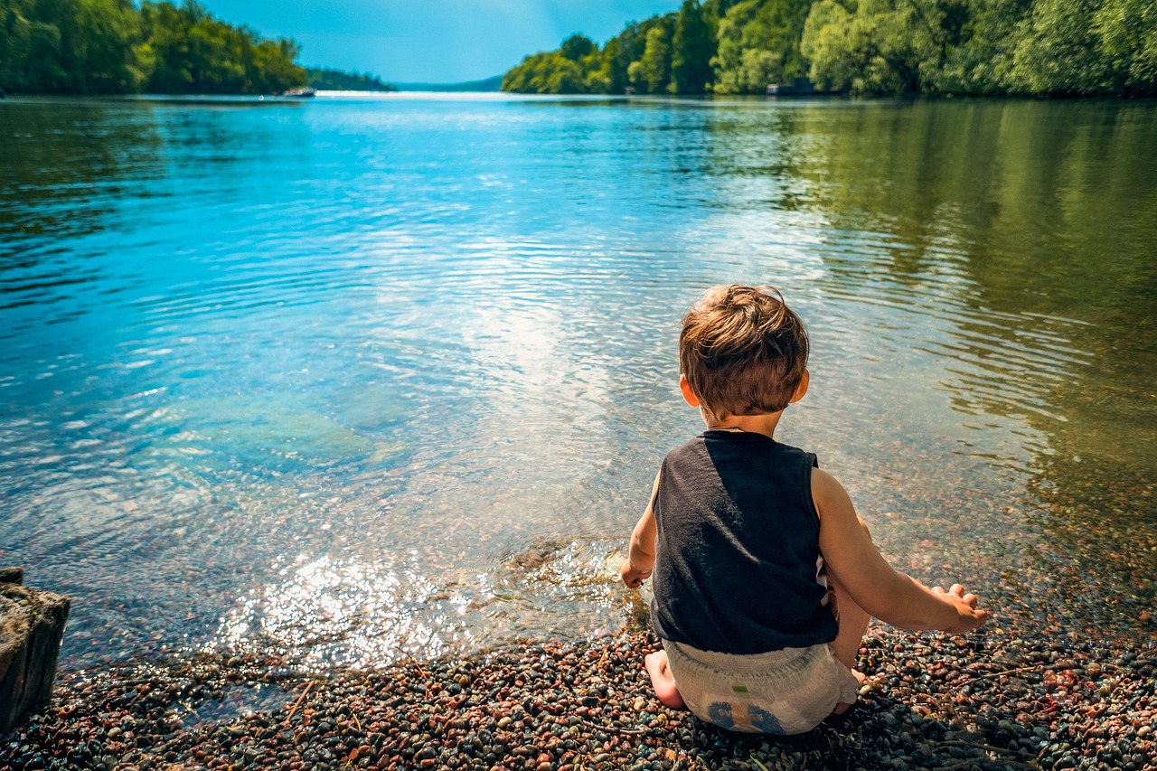 niño pequeño de espaldas en pañal y camiseta que está sentado en la orilla de un río o lago