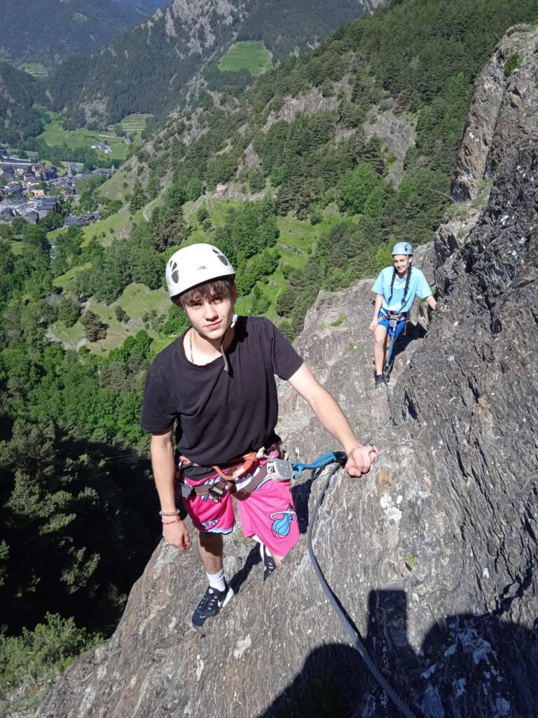 chico y chica escalando una montaña con cascos y arnés al fondo se ve un pueblo, montañas y árboles