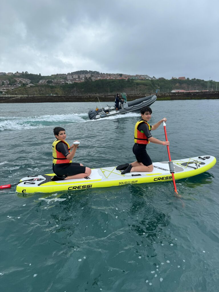 dos chavales en tabla de paddle surf, detrás de ellos una zodiac, al fondo un pueblo