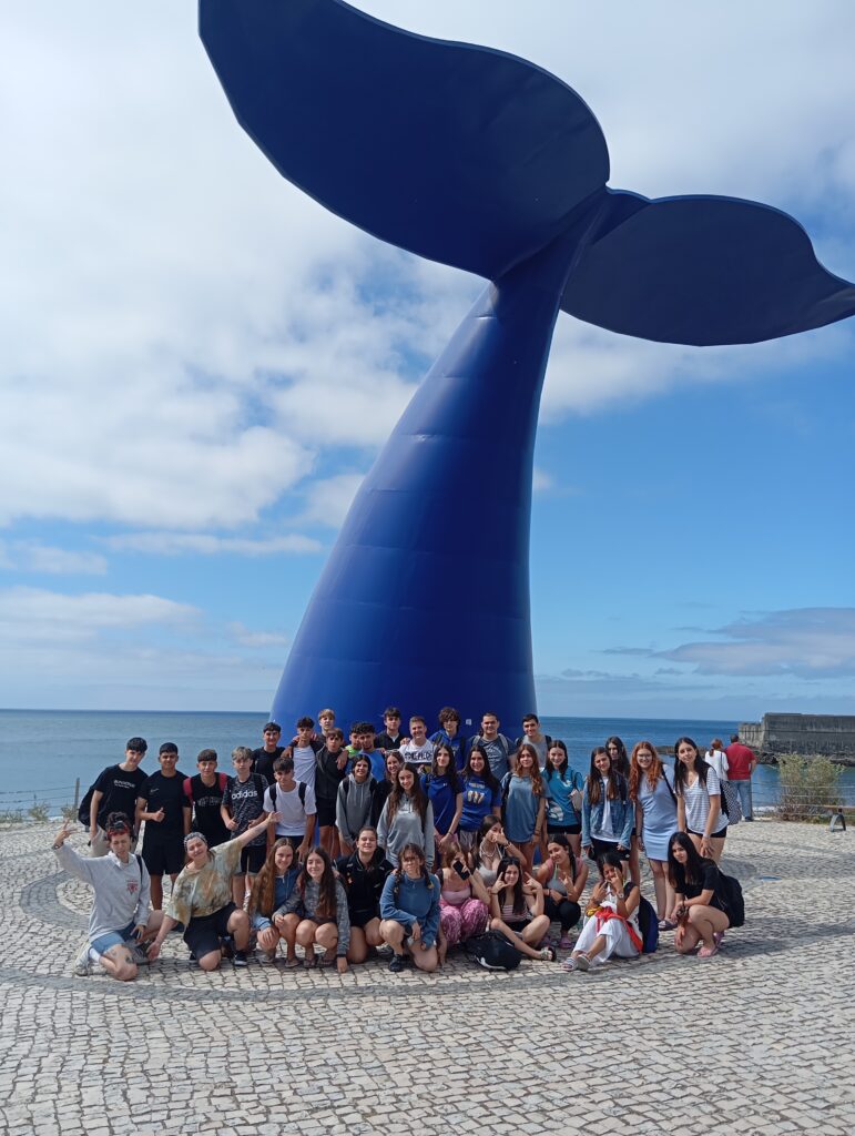 chicos y chicas delante de una escultura de la cola de una ballena, al fondo se ve el mar